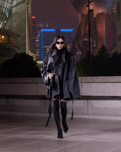 Woman in an oversized black leather cape and sunglasses walks through a night-time city plaza with illuminated skyscrapers in the background.
