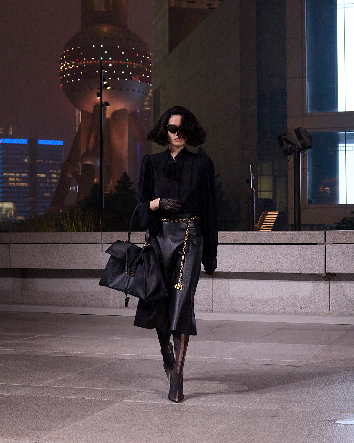 Person in black outfit walking at night in an urban plaza, wearing sunglasses and carrying a large black handbag.