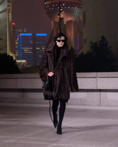 Woman walking at night in a long dark fur coat, black turtleneck, oversized sunglasses, and a chain-strap handbag against a lit urban skyline.