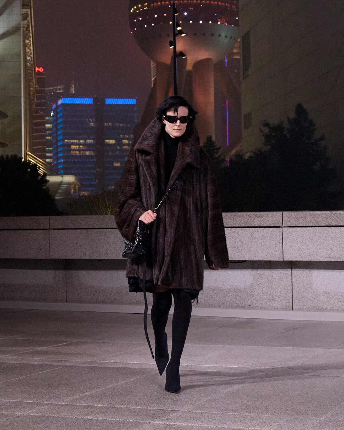 Woman walking at night in a long dark fur coat, black turtleneck, oversized sunglasses, and a chain-strap handbag against a lit urban skyline.