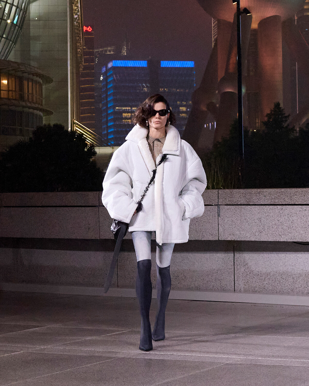 Woman in an oversized white coat and sunglasses posing on a city street at night, holding a black bag.