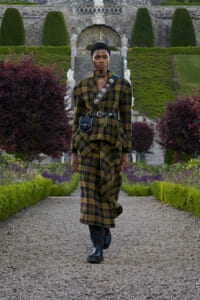 Model walking toward camera in a yellow‑black plaid coat with a studded crossbody bag, boots, and belt, along a gravel garden path with trimmed hedges and greenery behind.