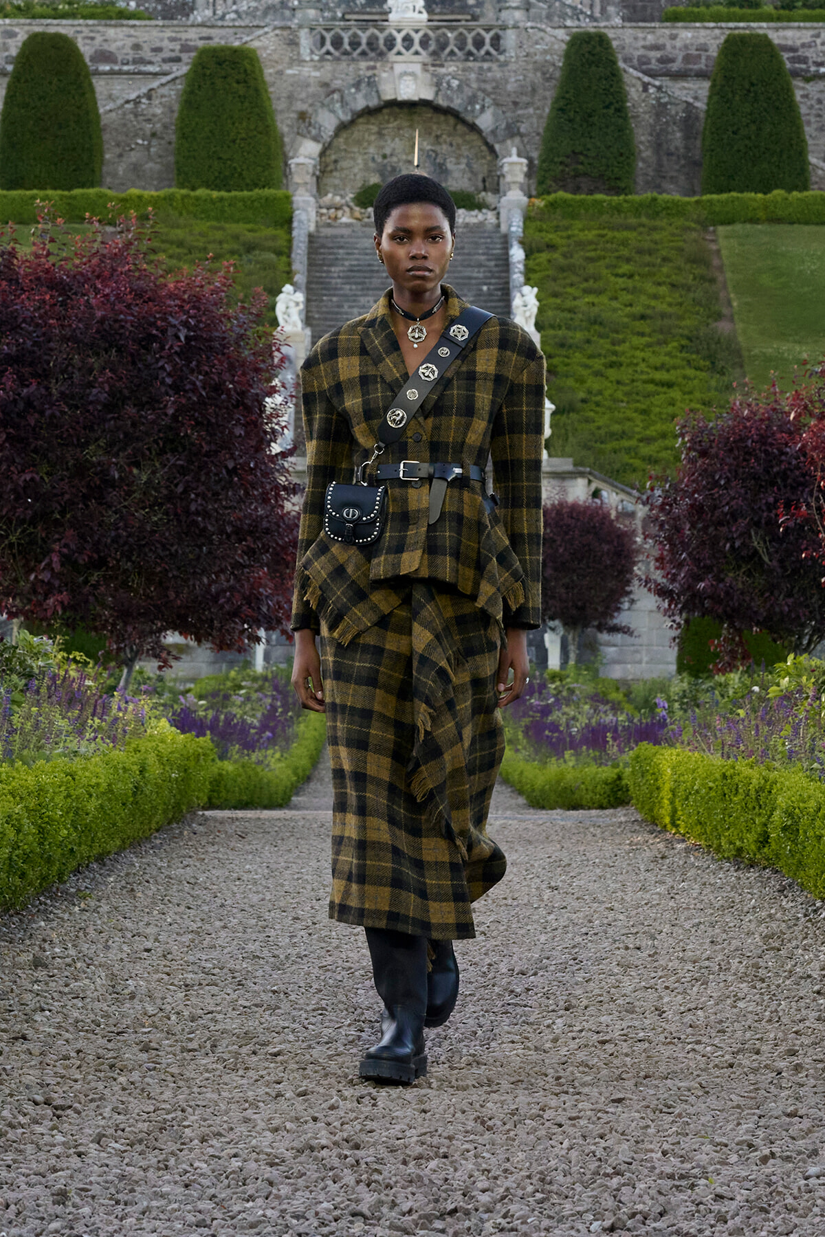 Model walking toward camera in a yellow‑black plaid coat with a studded crossbody bag, boots, and belt, along a gravel garden path with trimmed hedges and greenery behind.