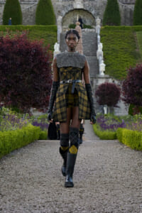 Model walking along a gravel path in a formal garden, wearing a yellow‑black plaid outfit with a studded capelet, belt, long gloves and knee‑high boots.