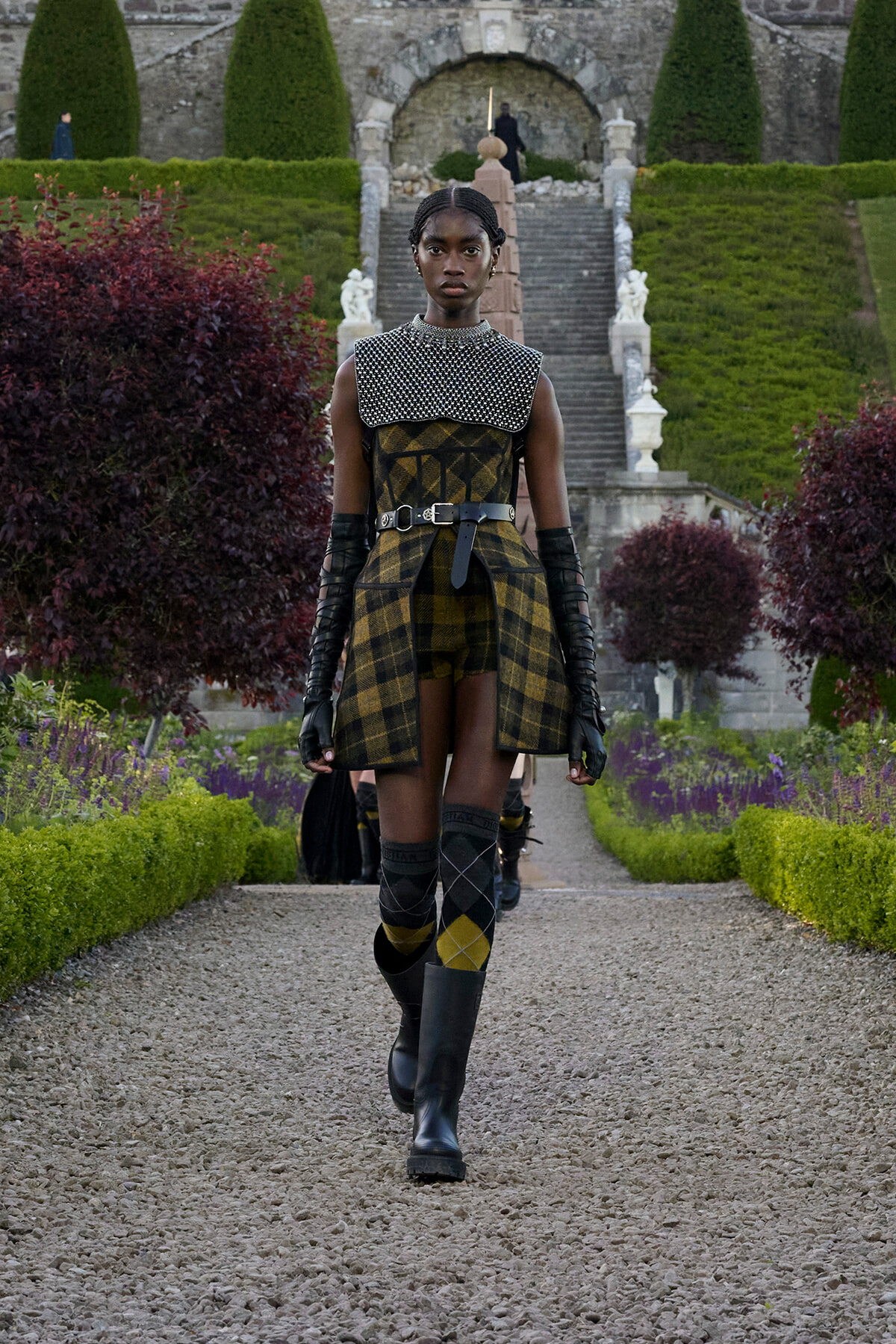 Model walking along a gravel path in a formal garden, wearing a yellow‑black plaid outfit with a studded capelet, belt, long gloves and knee‑high boots.