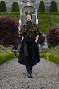 Model walking toward camera on a gravel garden path, wearing a black head wrap, mustard plaid cape, sheer black skirt and tall boots in a formal outdoor setting