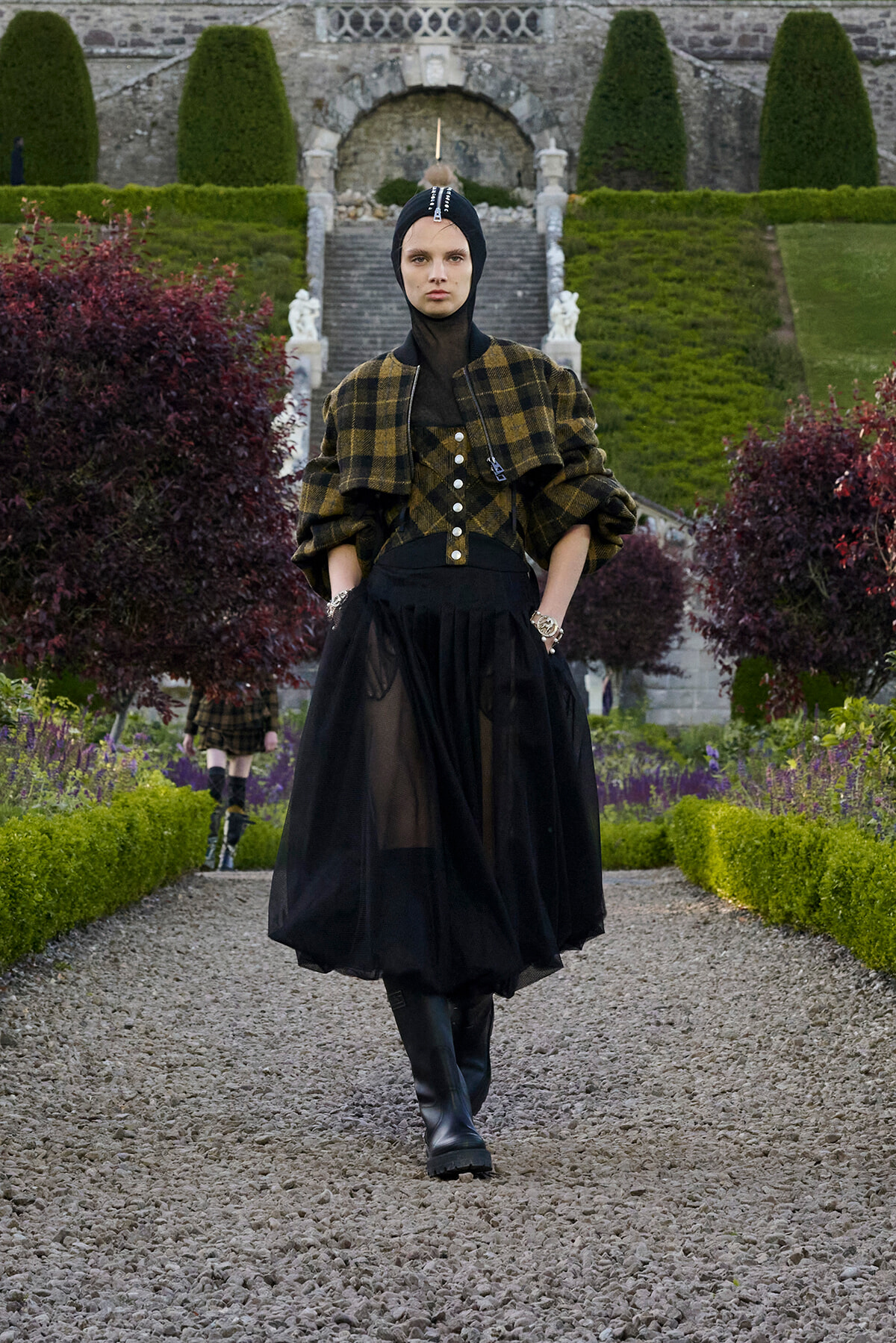 Model walking toward camera on a gravel garden path, wearing a black head wrap, mustard plaid cape, sheer black skirt and tall boots in a formal outdoor setting