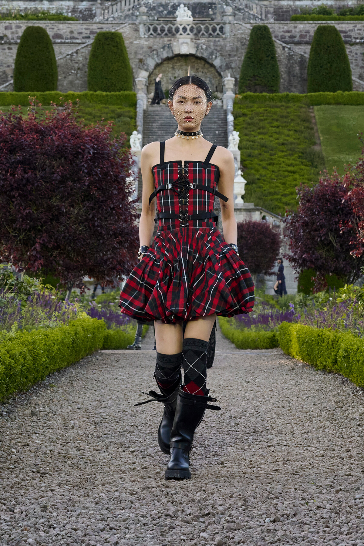 Model wearing a red-and-black plaid corset dress, choker, and tall boots walking down a gravel garden path with formal hedges and a stone staircase in the background.