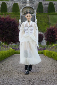 Model walking on a gravel path in a formal garden, wearing a white sheer skirt, white blouse, and a printed corset over the top, with black boots.