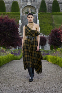 Model in a one-shoulder plaid dress with a black belt and chunky necklace walking down a gravel garden path in front of a stone staircase.