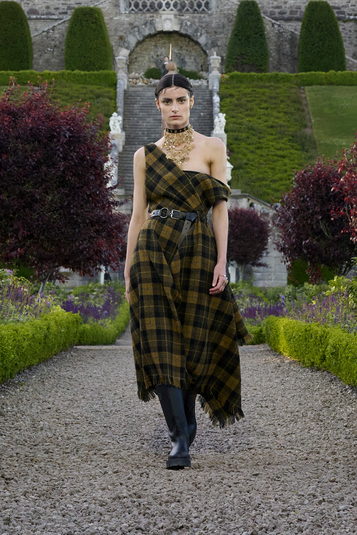 Model in a one-shoulder plaid dress with a black belt and chunky necklace walking down a gravel garden path in front of a stone staircase.