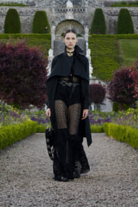 Model in a black Gothic lace outfit with cape and veil, walking a gravel path in a formal garden with trimmed hedges.