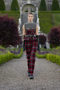 Female model walks a gravel garden runway in a red plaid jumpsuit with a studded silver collar and black gloves.