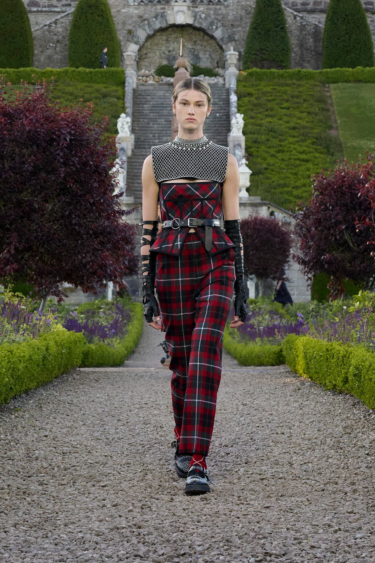 Female model walks a gravel garden runway in a red plaid jumpsuit with a studded silver collar and black gloves.