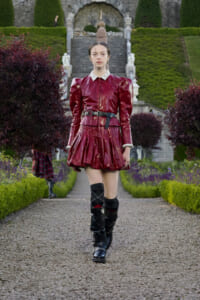 Model wearing a burgundy patent-leather dress with puff shoulders, lace collar, belt, and black knee-high boots in a formal garden setting.