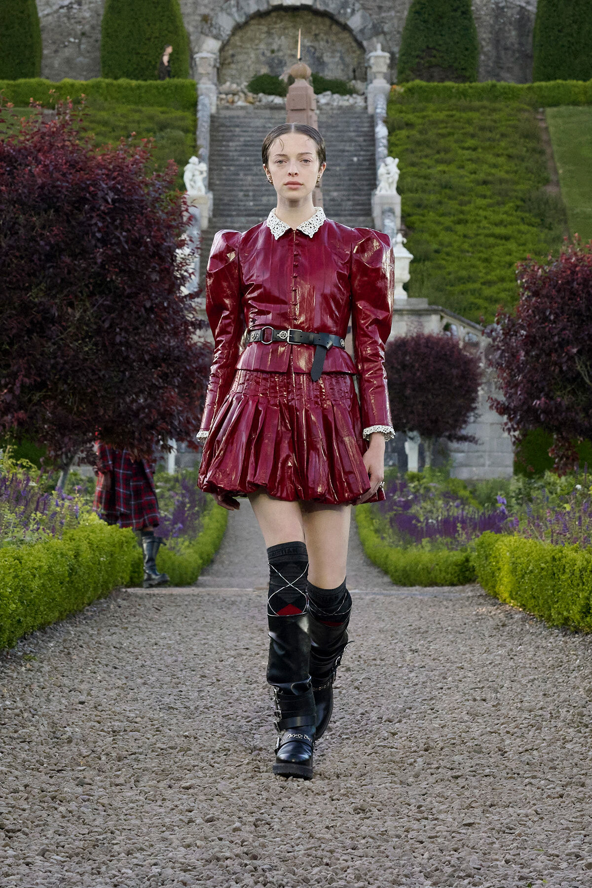 Model wearing a burgundy patent-leather dress with puff shoulders, lace collar, belt, and black knee-high boots in a formal garden setting.