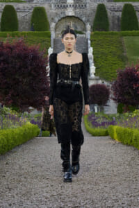 Model in a black lace dress with puff sleeves and a choker walks along a gravel garden path, with a stone staircase and manicured hedges in the background.