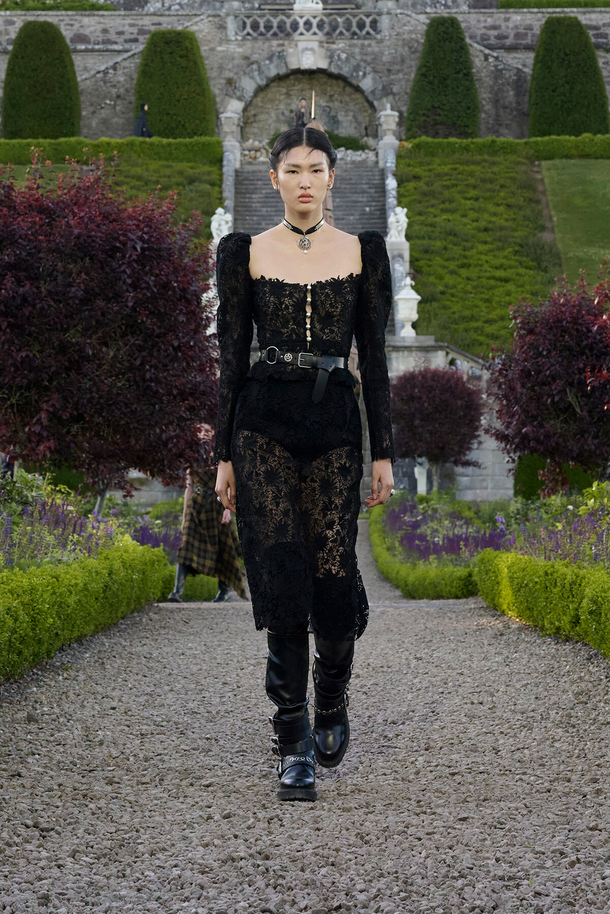Model in a black lace dress with puff sleeves and a choker walks along a gravel garden path, with a stone staircase and manicured hedges in the background.