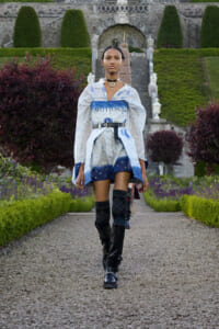 Model walking down a gravel garden path in a blue-toned outfit with 'EMOTIONAL' wording, belt, and knee-high socks, pair of black boots, formal garden backdrop.
