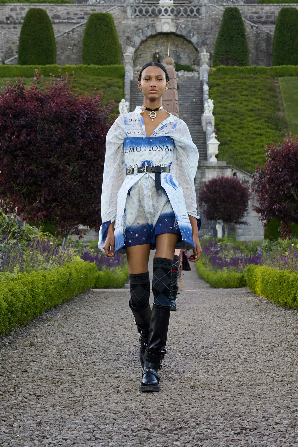 Model walking down a gravel garden path in a blue-toned outfit with 'EMOTIONAL' wording, belt, and knee-high socks, pair of black boots, formal garden backdrop.