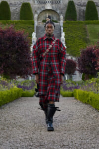 Person wearing a long red plaid coat over matching plaid clothing, with a decorative crossbody strap, walking along a gravel garden path in a formal stone garden setting with stairs in the background.