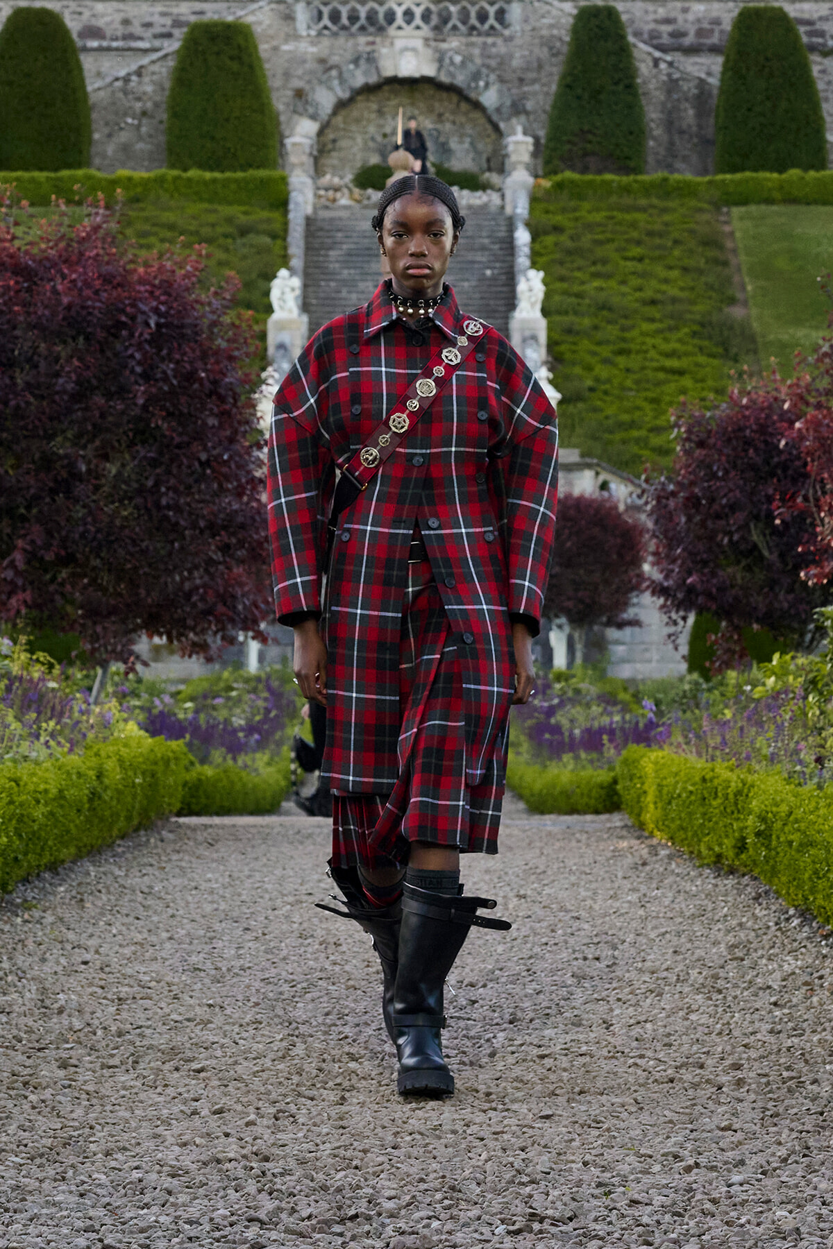 Person wearing a long red plaid coat over matching plaid clothing, with a decorative crossbody strap, walking along a gravel garden path in a formal stone garden setting with stairs in the background.
