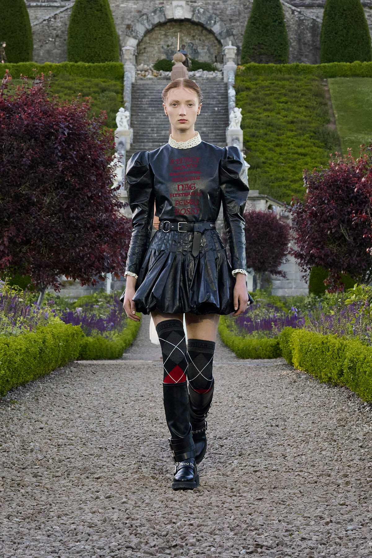 Model walking down a gravel garden path in a black leather dress with puff sleeves, belt, and argyle knee-high socks.
