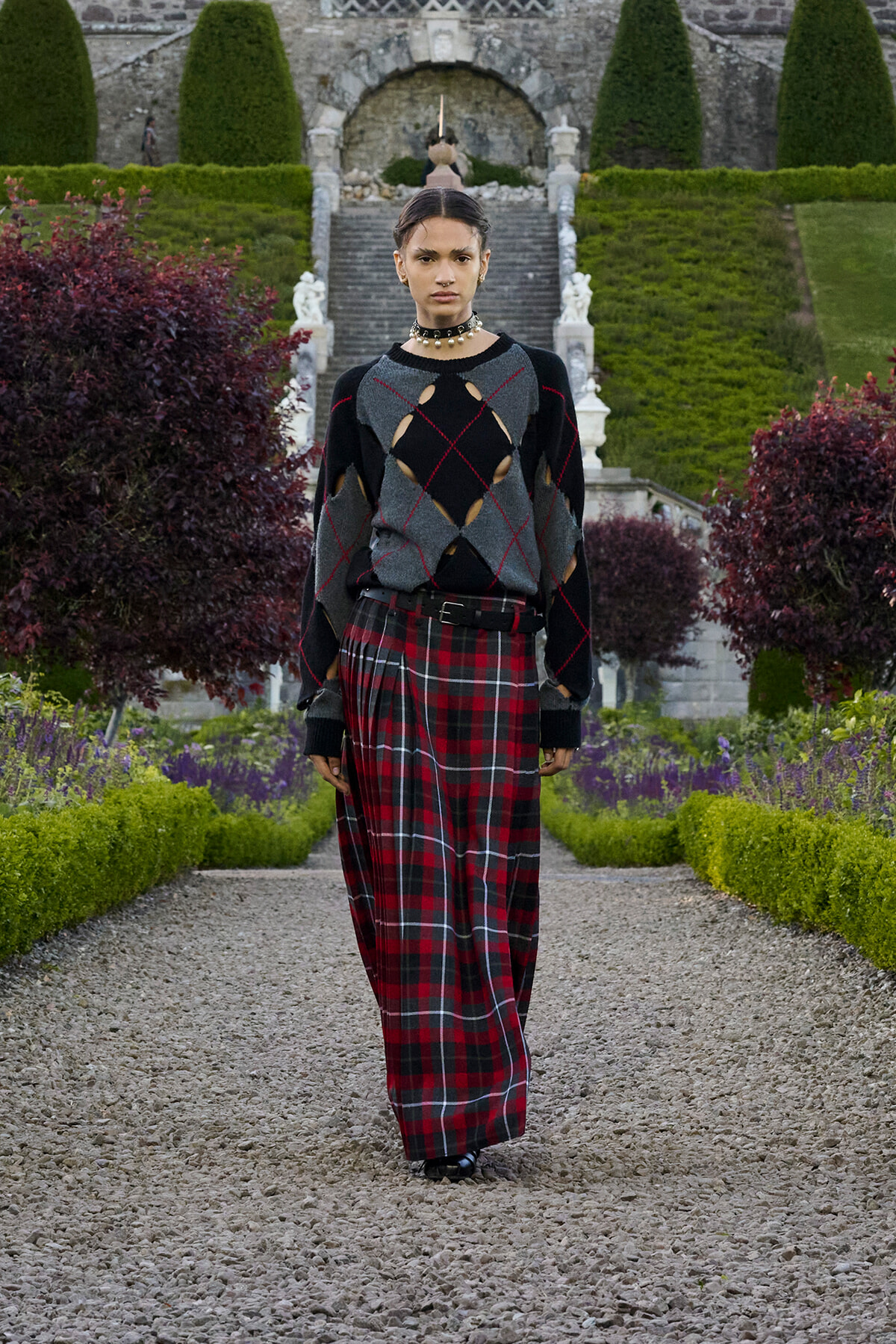 Model walking down a gravel garden path in a bold knit sweater with cutouts and a red plaid maxi skirt.