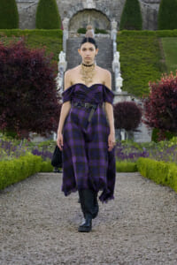 Model walking down a gravel garden path in a purple off-shoulder plaid jumpsuit with a black belt and layered gold necklace.