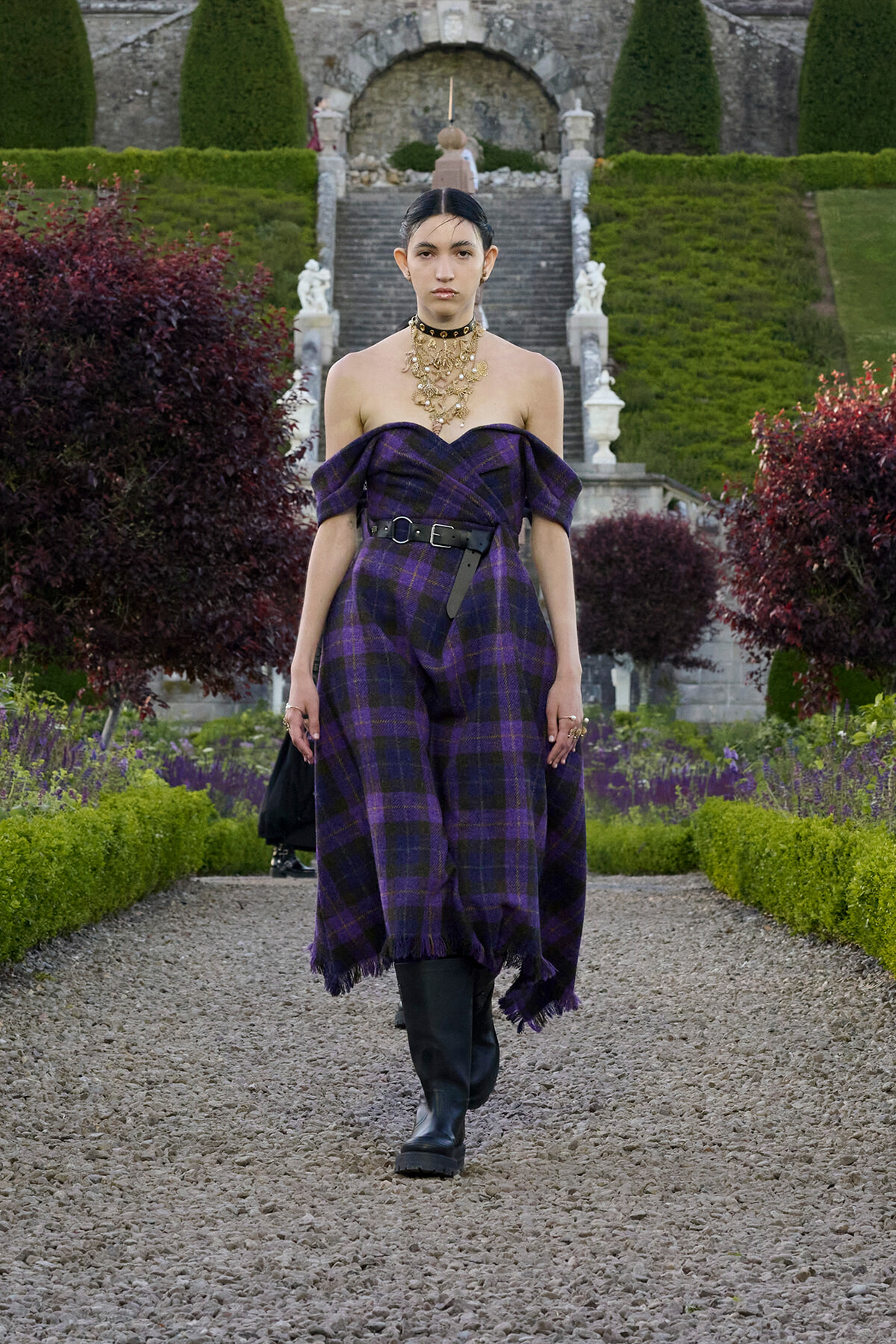 Model walking down a gravel garden path in a purple off-shoulder plaid jumpsuit with a black belt and layered gold necklace.