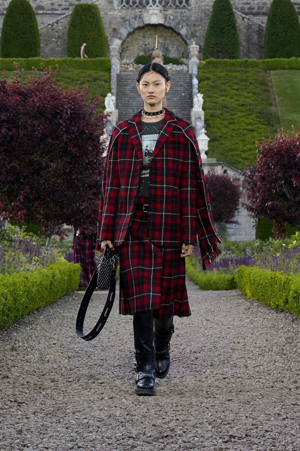 Person walking toward camera in a red plaid coat over black outfit, accessorized with a choker and studded bag in a formal garden setting.