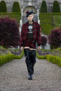 Person in red plaid jacket and matching shorts, black knee-high boots, walking along a gravel garden path.