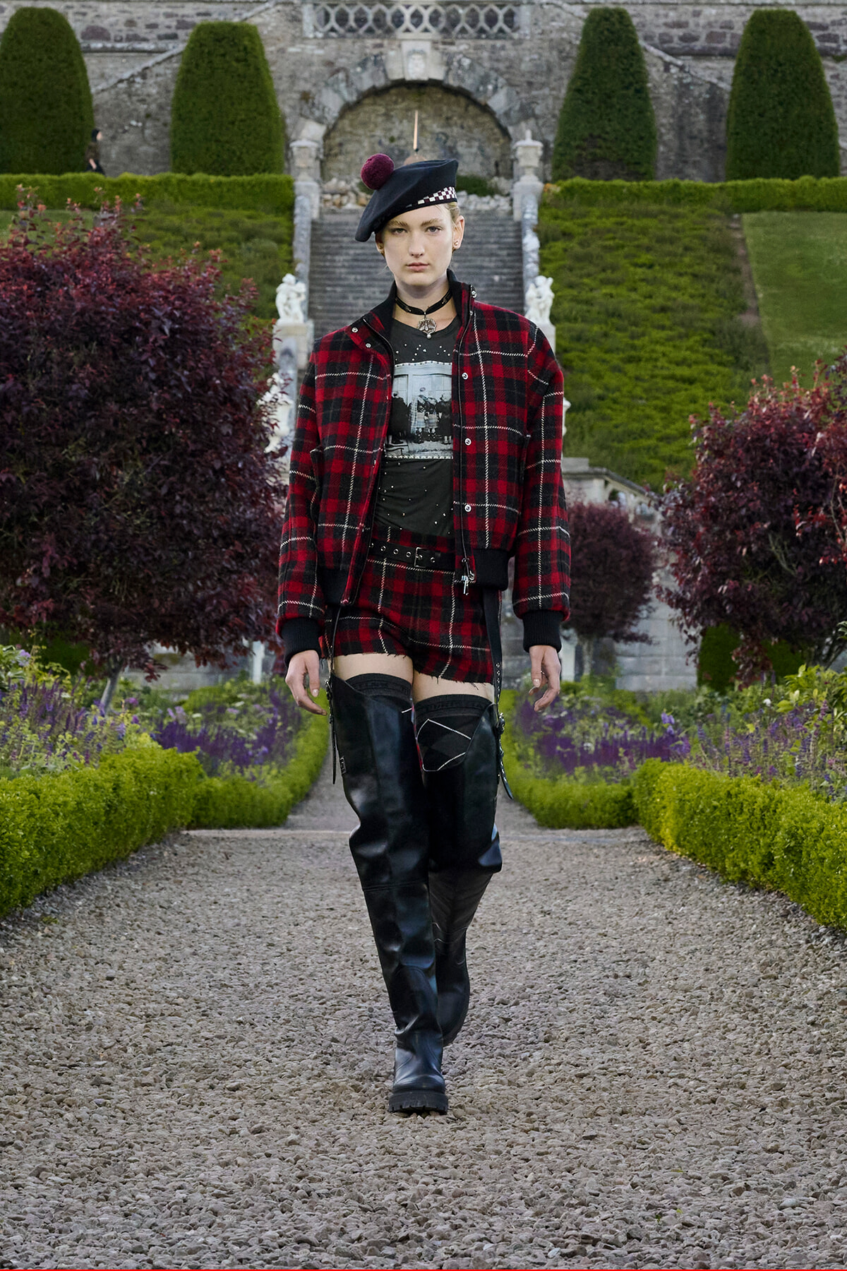 Person in red plaid jacket and matching shorts, black knee-high boots, walking along a gravel garden path.