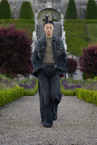 Model in studded dark denim suit walking along a gravel garden path with manicured hedges and stone stairs behind.