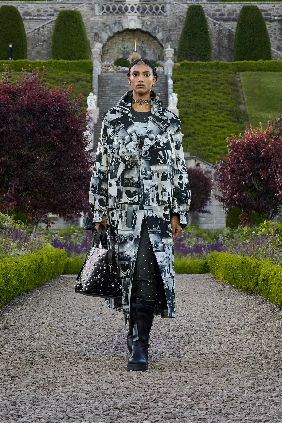 Model wearing a black-and-white photo-print trench coat walks down a gravel garden path, carrying a studded black handbag.