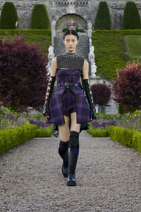 Model walking a gravel path in a purple plaid structured dress with a black belt, armored gloves, and knee-high boots in a formal garden.