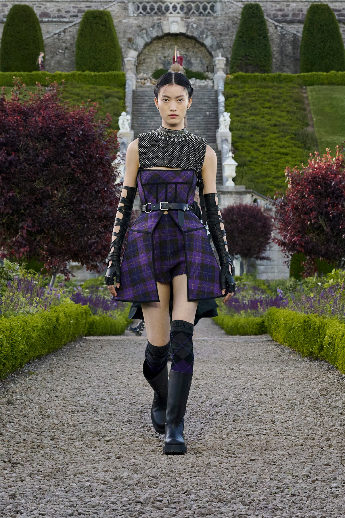 Model walking a gravel path in a purple plaid structured dress with a black belt, armored gloves, and knee-high boots in a formal garden.
