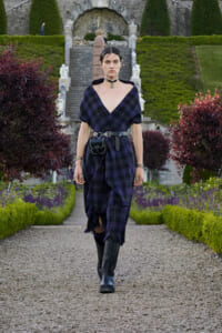 Person walking toward the camera along a gravel garden path, wearing a dark purple plaid dress with a black belt and knee-high boots; manicured hedges and stone stairs visible in the background.