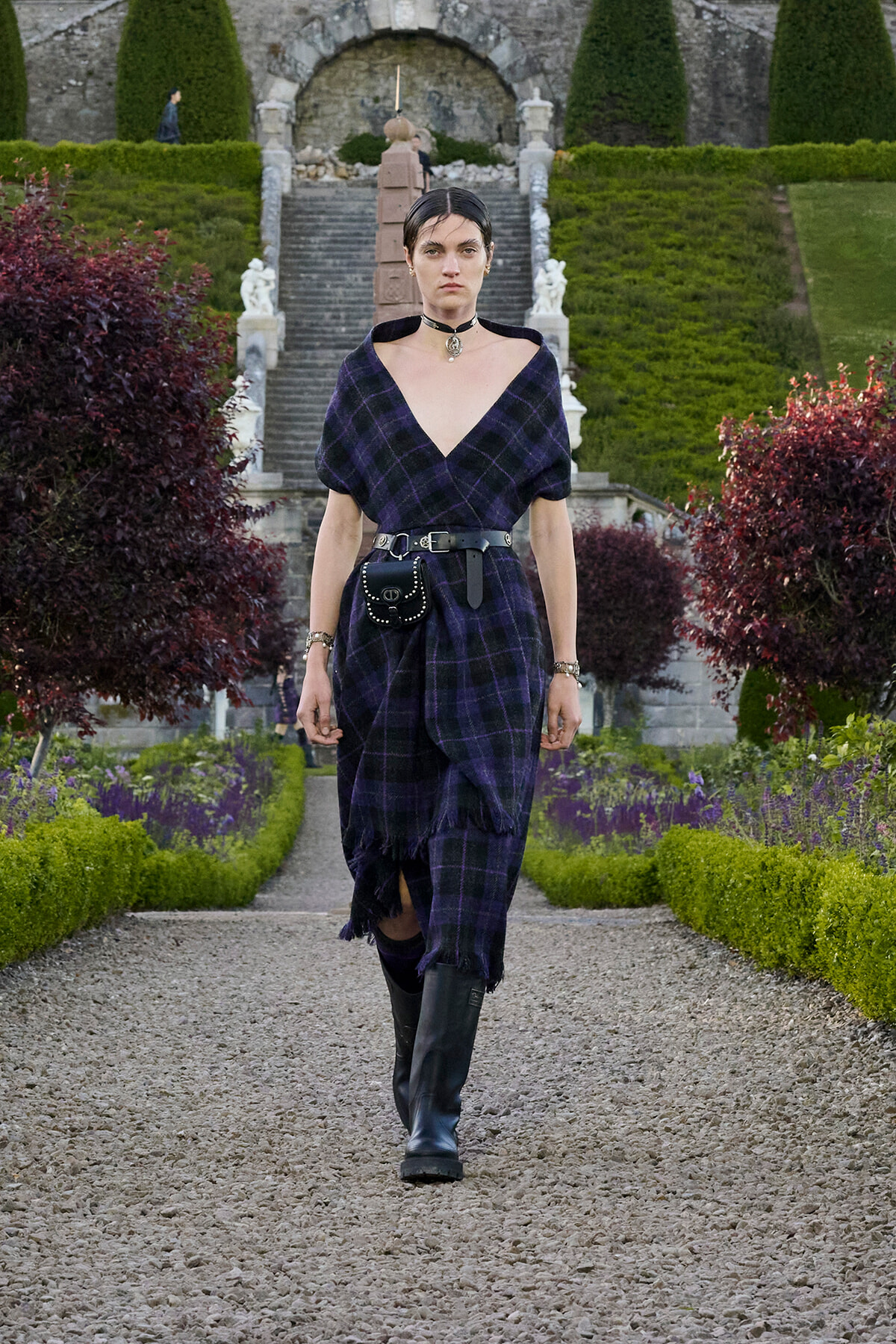 Person walking toward the camera along a gravel garden path, wearing a dark purple plaid dress with a black belt and knee-high boots; manicured hedges and stone stairs visible in the background.