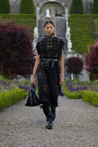 Model walking down a gravel path in a formal garden, wearing a sheer black fringe top, black pants, boots, and a studded belt bag. Holding a black handbag.