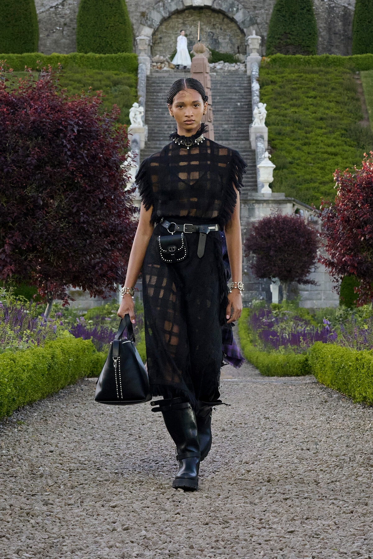 Model walking down a gravel path in a formal garden, wearing a sheer black fringe top, black pants, boots, and a studded belt bag. Holding a black handbag.