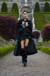 Model walking toward the camera in a black Gothic outfit with a corset, patches, and cape, in a formal garden background.