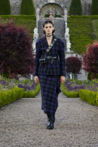 Person wearing a dark purple plaid suit with a belt bag, walking down a gravel garden path.