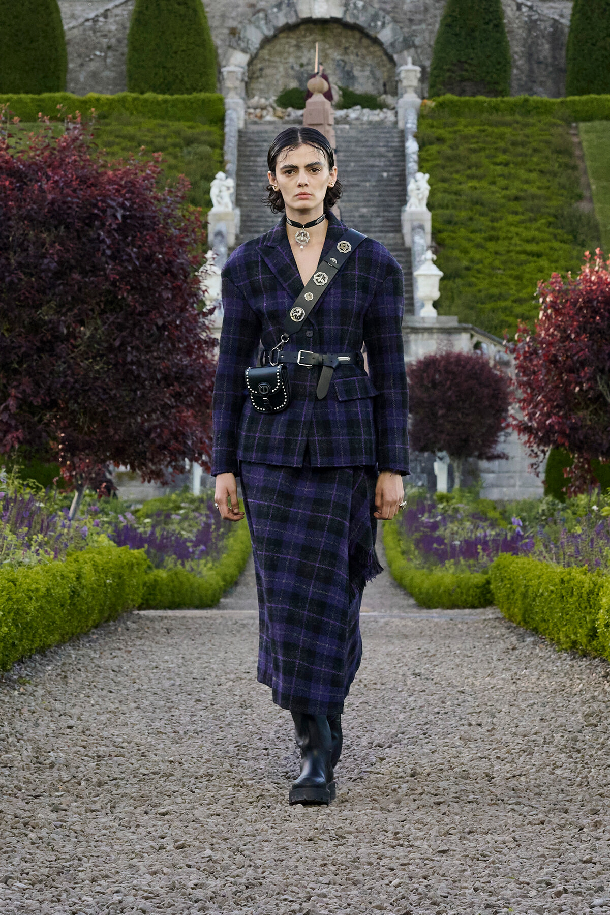 Person wearing a dark purple plaid suit with a belt bag, walking down a gravel garden path.
