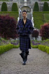 Person walking toward camera in a dark purple fur jacket and matching plaid skirt, black boots, wearing a choker, in a formal garden with trimmed hedges and stone steps in the background.
