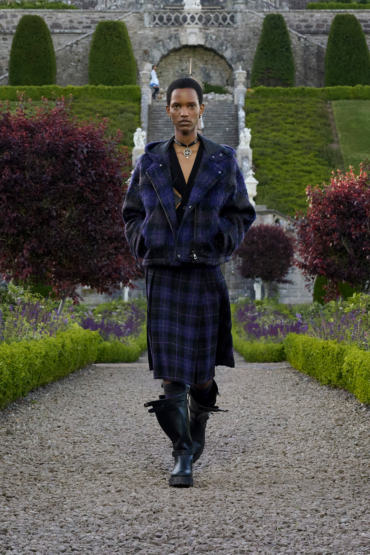 Person walking toward camera in a dark purple fur jacket and matching plaid skirt, black boots, wearing a choker, in a formal garden with trimmed hedges and stone steps in the background.
