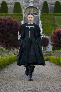 Model walking toward the camera in a dramatic black, layered outfit with boots in a formal garden with stone steps and manicured hedges behind.