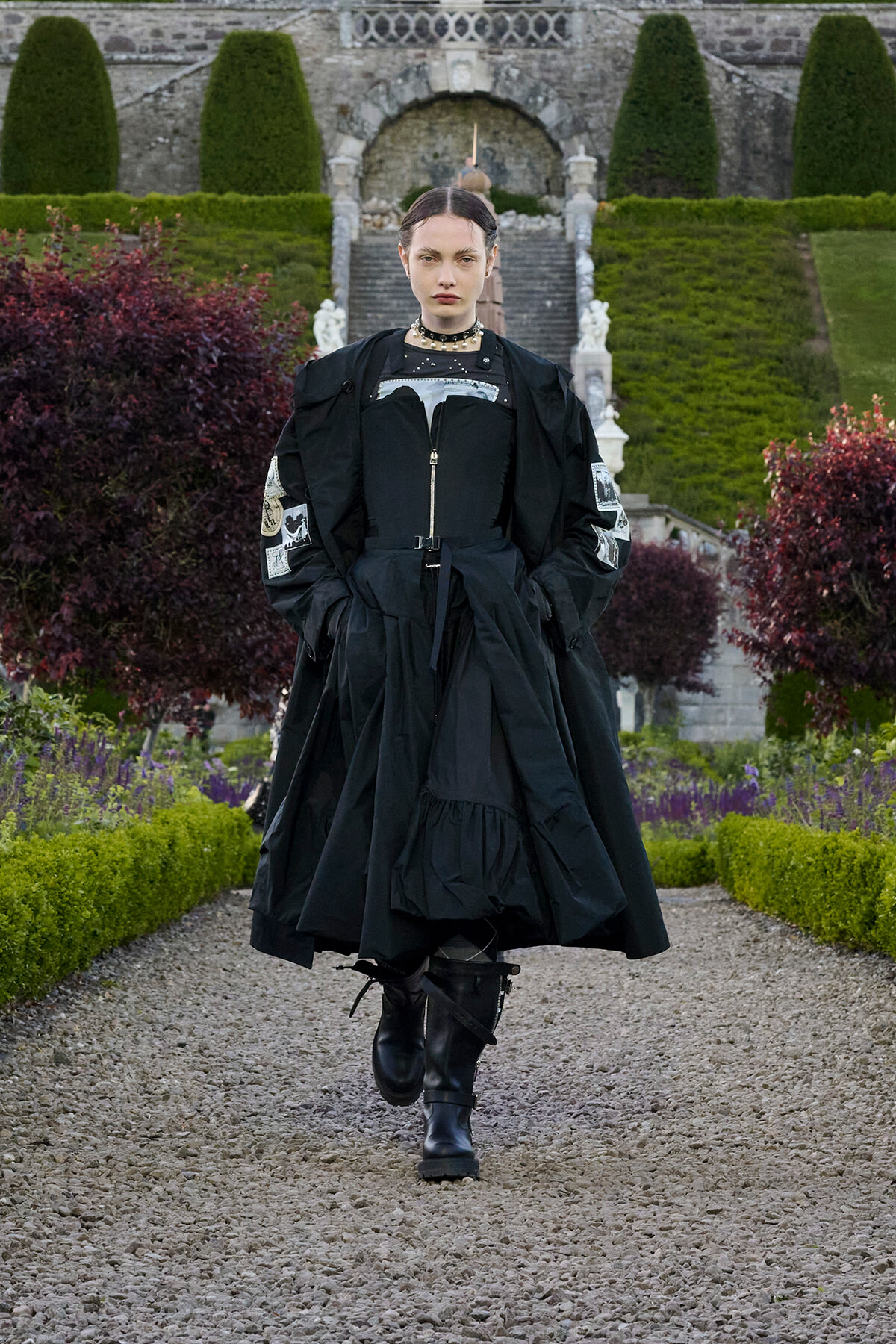 Model walking toward the camera in a dramatic black, layered outfit with boots in a formal garden with stone steps and manicured hedges behind.