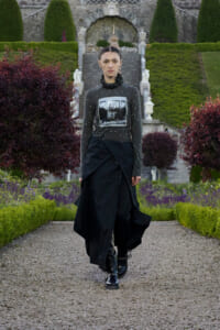 Model walking a gravel garden path in a dark, pearl-studded top with a photo print and a long black layered skirt with boots, in a formal garden setting.