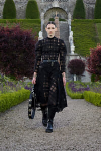 Model in a black sheer lace dress with a wide belt and knee-high boots walks along a gravel garden path, carrying a studded black bag.